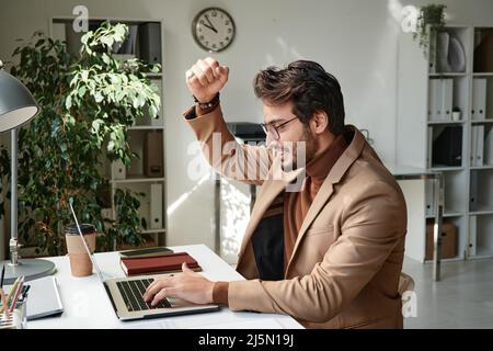 Lächelnder, gutaussehender Manager aus dem Nahen Osten in Brillen, der mit Ja-Gesten und einem Laptop unterwegs war, während er ein Ergebnis erzielt hat Stockfoto