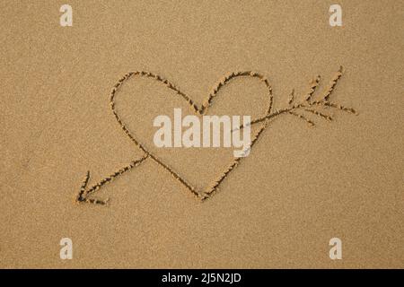 Ein von einem Pfeil durchbohrtes Herz wird auf den weichen Strandsand gezogen. Stockfoto