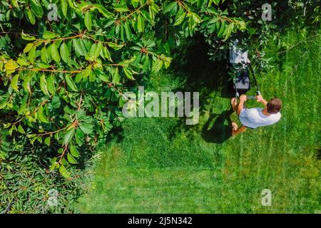 Gärtner schiebt Rasenmäher zum Schneiden von grünem Gras im Garten im Sommer. Luftaufnahme. Stockfoto