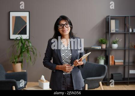 Horizontales mittleres Porträt einer professionellen Psychologin, die eine Brille trägt und ein digitales Tablet in ihrem Büro hält, das lächelt und die Kamera anlächelt Stockfoto