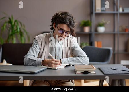 Horizontales, mittleres Porträt eines jungen erwachsenen Mannes, der ein bequemes, legeres Outfit trägt und am Schreibtisch sitzende Brillen hat, die etwas im Notizbuch schreiben Stockfoto