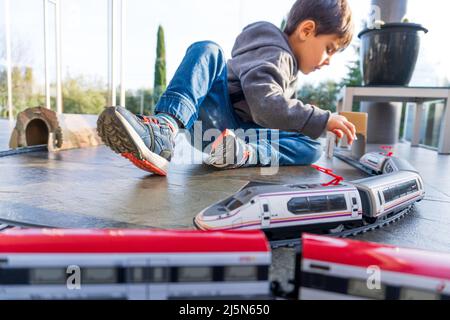 VJunges Kind, das mit Spielzeugzügen auf dem Boden spielt. Spielzeugzug ähnlich dem spanischen Zug Cercanias Renfe und dem Hochgeschwindigkeitszug Ave Stockfoto