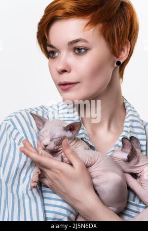 Schöne rothaarige junge Frau umarmt Sphynx Cat und schaut weg. Studio auf weißem Hintergrund aufgenommen. Portrait von Hipster mit kurzen Haaren Stockfoto