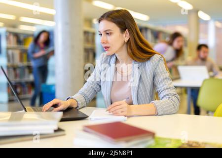 Junge Frau, die mit Büchern und Laptop an einem Projekt in der Bibliothek arbeitet Stockfoto