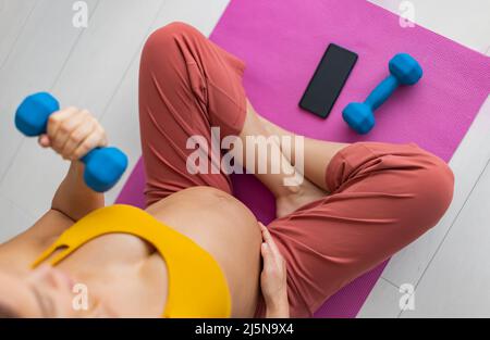 Schwanger Frau trainieren zu Hause Training mit Online-Fitness-Handy-App heben freies Gewicht für Arm-Training Stockfoto