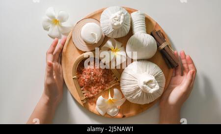 Draufsicht, weibliche Hände halten einen Korb mit Spa-Massageprodukten auf weißem Hintergrund. Massagebälle, Himalaya-Salz, Duftkerze, Kräuter und Frangipan Stockfoto