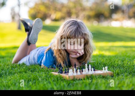 Konzentrierter Junge, der eine Schachstrategie entwickelt und ein Schachbrettspiel spielt. Netter kleiner Junge, der Schach spielt und im Sommerpark auf Gras liegt. Stockfoto