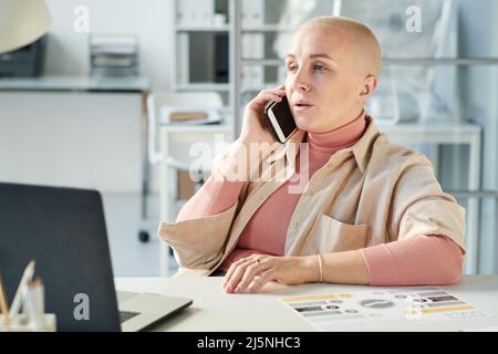 Selbstbewusste kahle kaukasische Frau im rosa Rollkragen sitzt vor dem Laptop und diskutiert Projekt am Telefon Stockfoto