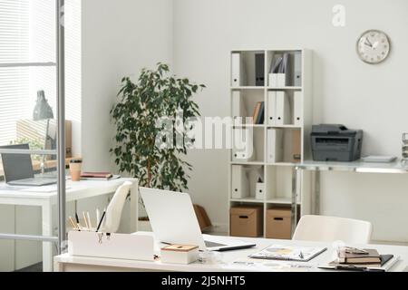 Leerer moderner Büroarbeitsplatz mit Laptops, Verbrauchsmaterialien und Papieren auf Tischen, Regalen mit Ordner und Drucker im Hintergrund Stockfoto