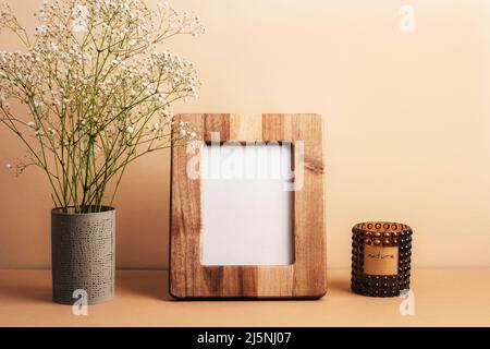 Leerer Bilderrahmen, Duftkerze und Gypsophila-Blüten in Vase auf beigem Tisch. Vorderansicht, Modell. Stockfoto