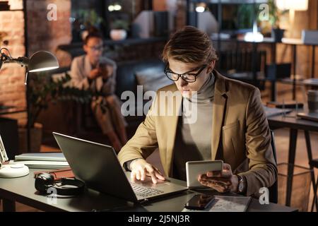 Serious jungen kaukasischen Geschäftsmann in Jacke sitzt am Schreibtisch unter Lampe Licht und Analyse von Online-Daten auf Geräten Stockfoto