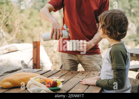 Vater Vater gießt heißen Kaffee-Tee aus Thermoskannen in den Becher bei einem Familienpicknick in den Bergen. Das Kind der Kinderschule sieht zu, wie sein Vater die füllt Stockfoto