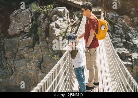 Vater und Sohn blicken beim Wandern weit von der Seilbrücke in die Berge. Lässig gekleidete Touristen Kinderschultjunge und sein Vater mit gelb Stockfoto