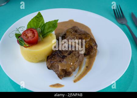 Fleischbällchen mit Kartoffelpüree auf einem weißen Teller. Fleisch in saurer Rahmsoße. Tomaten und Basilikum. Essen im Restaurant. Mittagspause. Medaillons. Stockfoto