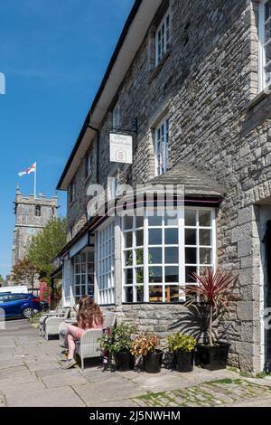 Junge Frau, die ein Taschenbuch vor einem Bed & Breakfast-Restaurant und einer Brasserie in Corfe Castle Village, Dorset, England, GB, liest Stockfoto