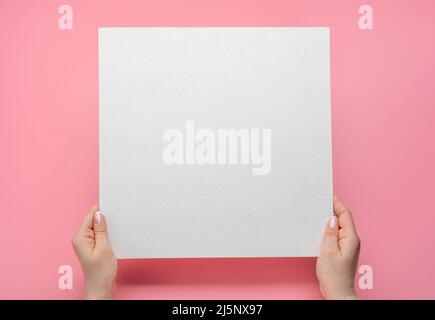 Viereckiges Mockup-Papier. Über dem Kopf gedrehte Aufnahme von Frauenhänden mit leerem Papierblatt auf einem rosafarbenen Tisch. Menschliche Hände halten eine weiße Werbekarte mit Mockup. Isoliert ein Stockfoto