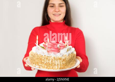 Eine brünette Frau in roten Kleidern hält einen großen weißen Kuchen mit Kerzen, macht einen Wunsch Stockfoto