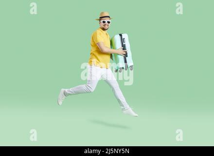 Fröhlicher männlicher Tourist in Sommerkleidung, der mit Koffer auf hellgrünem Hintergrund läuft. Stockfoto