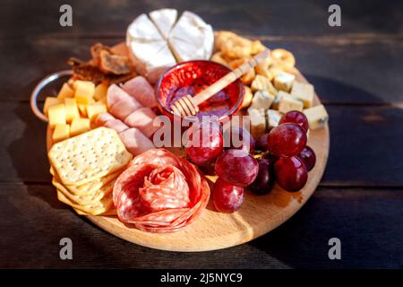 Rundes Schneidebrett mit verschiedenen Käsesorten, Wurstrosette, Schinken, Cracker, Trauben und Feigen mit Honig. Aufschnitt und Käseplatte auf dem Tter Stockfoto