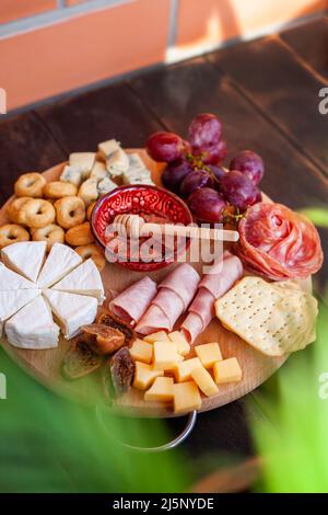 Rundes Schneidebrett mit verschiedenen Käsesorten, Wurstrosette, Schinken, Cracker, Trauben und Feigen mit Honig. Aufschnitt und Käseplatte auf dem Tter Stockfoto