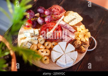 Rundes Schneidebrett mit verschiedenen Käsesorten, Wurstrosette, Schinken, Cracker, Trauben und Feigen mit Honig. Aufschnitt und Käseplatte auf dem Tter Stockfoto