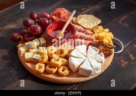 Rundes Schneidebrett mit verschiedenen Käsesorten, Wurstrosette, Schinken, Cracker, Trauben und Feigen mit Honig. Aufschnitt und Käseplatte auf dem Tter Stockfoto