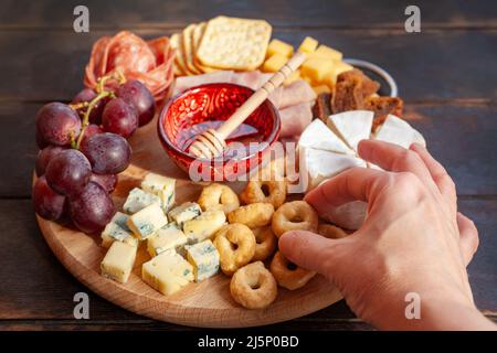 Die Hand der Frau nimmt einen kleinen salzigen Plätzchen vom Vorspeisenbrett mit verschiedenen Käsesorten, Fleisch, Wurstrosette, Trauben und Keksen. Charcuterie und Chee Stockfoto