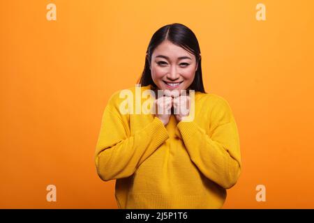 Überglücklich wunderschöne asiatische Frau lächelt herzlich, während sie einen lebhaften Pullover auf orangefarbenem Hintergrund trägt. Aufgeregt freundlich positive Mädchen hält die Arme an der Brust, während glücklich und fröhlich. Stockfoto