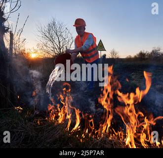 Feuerwehrmann-Ökologe, der das Feuer auf dem Feld mit dem Abendhimmel im Hintergrund bekämpft. Mann hält Eimer und gießt Wasser auf brennendes trockenes Gras in der Nähe des gelben Dreiecks mit Totenkopf und Kreuzknochen Warnschild. Stockfoto