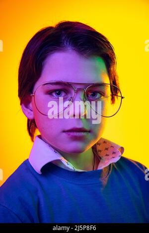 Nahaufnahme eines Jungen, eines Kindes in blauem Pullover, Hemd und Brille, isoliert auf gelbem Studiohintergrund in Neonlicht Stockfoto