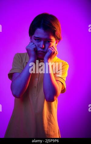 Porträt eines Jungen, Kind, das Wangen mit Händen mit gelangweilter unangenehmer Expression auf violettem Hintergrund in Neon isoliert Stockfoto