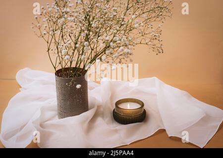 Kerze und Gypsophila Blumen in Vase auf weißer Tischdecke auf beigem Tisch. Stillleben. Stockfoto