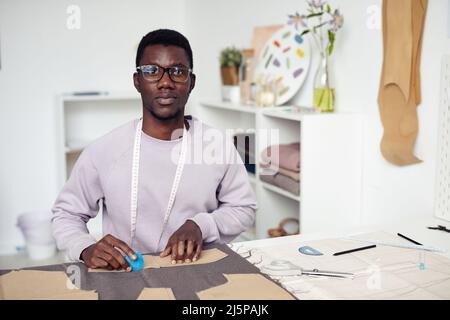 Porträt eines jungen schwarzen Schneiders, der in seinem Atelier an einem großen Tisch sitzt und Kleidungsdetails umreißt und ausschneidet Stockfoto