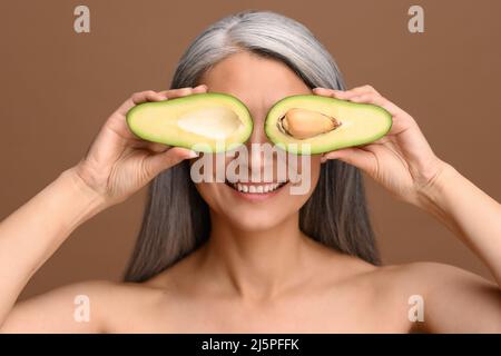 Schöne asiatische 50s reife grauhaarige Frau, die die Augen mit Stücken frischer Avocado bedeckt, die auf Braun isoliert sind. Attraktive reife Dame empfiehlt ölreiche Lebensmittel. Gesundes Essen und Lifestyle-Konzept Stockfoto