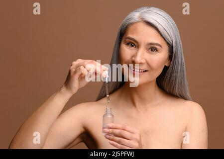 Porträt einer schönen asiatischen Frau mittleren Alters mit grauen Haaren hält Flasche mit kosmetischem Öl oder Verjüngungserum isoliert auf beige braunen Hintergrund, 50s weibliche Werbung Kosmetik-Produkt Stockfoto