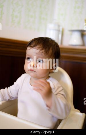 Weibliches kaukasisches Baby, das während ihres Geburtstags auf ihrem Stuhl sitzt und Schlagsahne auf ihrer Nase hat Stockfoto