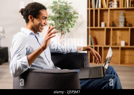 Seitenansicht des multirassischen jungen Mannes nutzt einen Laptop für einen Videoanruf zu Hause, winkt er Hallo zur Webcam. Videokommunikation, Zoom Stockfoto