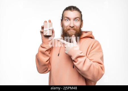 Scandinavian handsome Mann hält Geschenk und zeigt isoliert auf grauem Hintergrund zu präsentieren. Kerl mit Ingwer-Frisur und Bart in einem Sweatshirt. Konzept Stockfoto