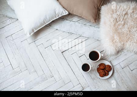Frühstück am Morgen. Zwei weiße Tassen Kaffee und Schokoladenkekse stehen auf einem hellen Holzparkett und Kissen liegen in der Nähe. Gemütlichkeit, Tag Stockfoto