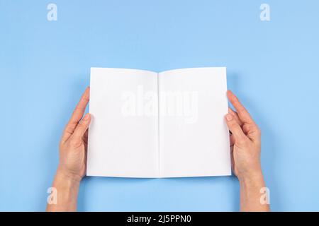 Frau Hände mit gefaltetem leerem Papierbogen, leerem Booklet, doppelseitigem Buch, Magazin auf hellblauem Hintergrund. Mustervorlage. Draufsicht Stockfoto