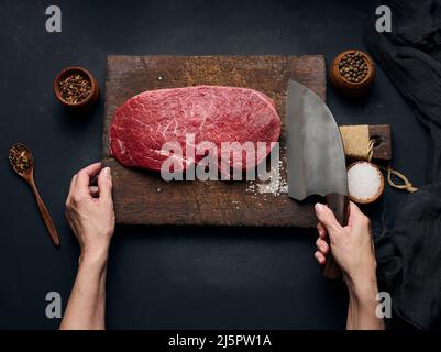 Roh-Rinderfilet liegt auf einem Holzschneidebrett und Gewürze zum Kochen auf einem schwarzen Tisch, Draufsicht. Die Hand einer Frau hält ein großes Küchenmesser in sich Stockfoto