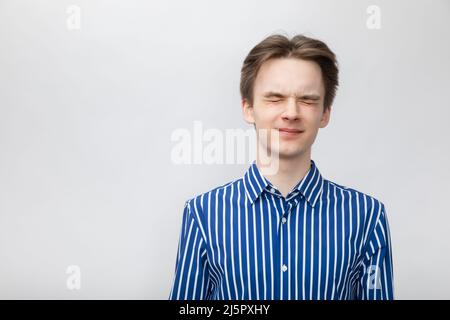 Porträt eines jungen Mannes mit blau-weiß gestreiftem Knopfhemd schließt seine Augen fest. Studio auf grauem Hintergrund aufgenommen Stockfoto