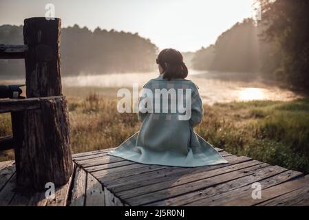 Rückansicht einer Frau, die auf der Holzbrücke sitzt und den Nebel über dem See in Pang Oung, Provinz Mae Hong Son, betrachtet. Stockfoto