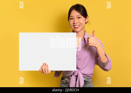 Schöne junge asiatische Frau mit leerem weißen Papierbanner mit Mockup und zeigt Daumen nach oben Geste auf gelbem Hintergrund Stockfoto