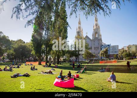 USA, Washington Square Park in San Francisco Menschen liegen auf dem Rasen mit der St. Peter und Paul Kirche im Hintergrund und einer Frau auf einem Fatboy-Wor Stockfoto