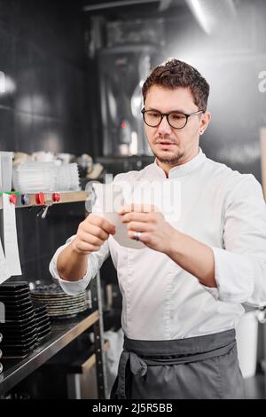 Professioneller Koch in der Schürze hält Papier in den Händen und prüft Reihenfolge des Gastes vor dem Kochen Stockfoto