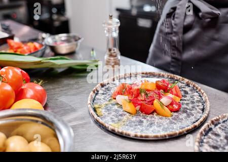Nahaufnahme des Gerichts mit frischem Gemüsesalat auf dem Teller auf dem Tisch, wobei der Koch Gewürze hinzufügte Stockfoto