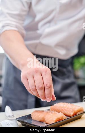 Nahaufnahme des Küchenchefs, der Lachsfisch salzt, um das Gericht am Tisch in der Küche zuzubereiten Stockfoto