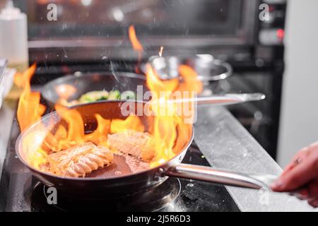 Nahaufnahme des Kochs, der die Pfanne über dem Herd hält, und Braten von Fisch auf offenem Feuer in der Küche Stockfoto