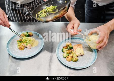 Nahaufnahme des Küchenchefs, der das Gericht auf Teller legt, während sein Assistent am Tisch die Sauce auf das Gericht gießt Stockfoto